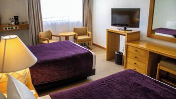 A hotel room with two beds, a desk with a TV, a chair, a lamp, and a window with curtains.