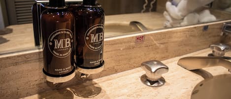 Casa de banho com lavatório, espelho e duas embalagens de gel de mãos e corpo com a etiqueta 'MB'.