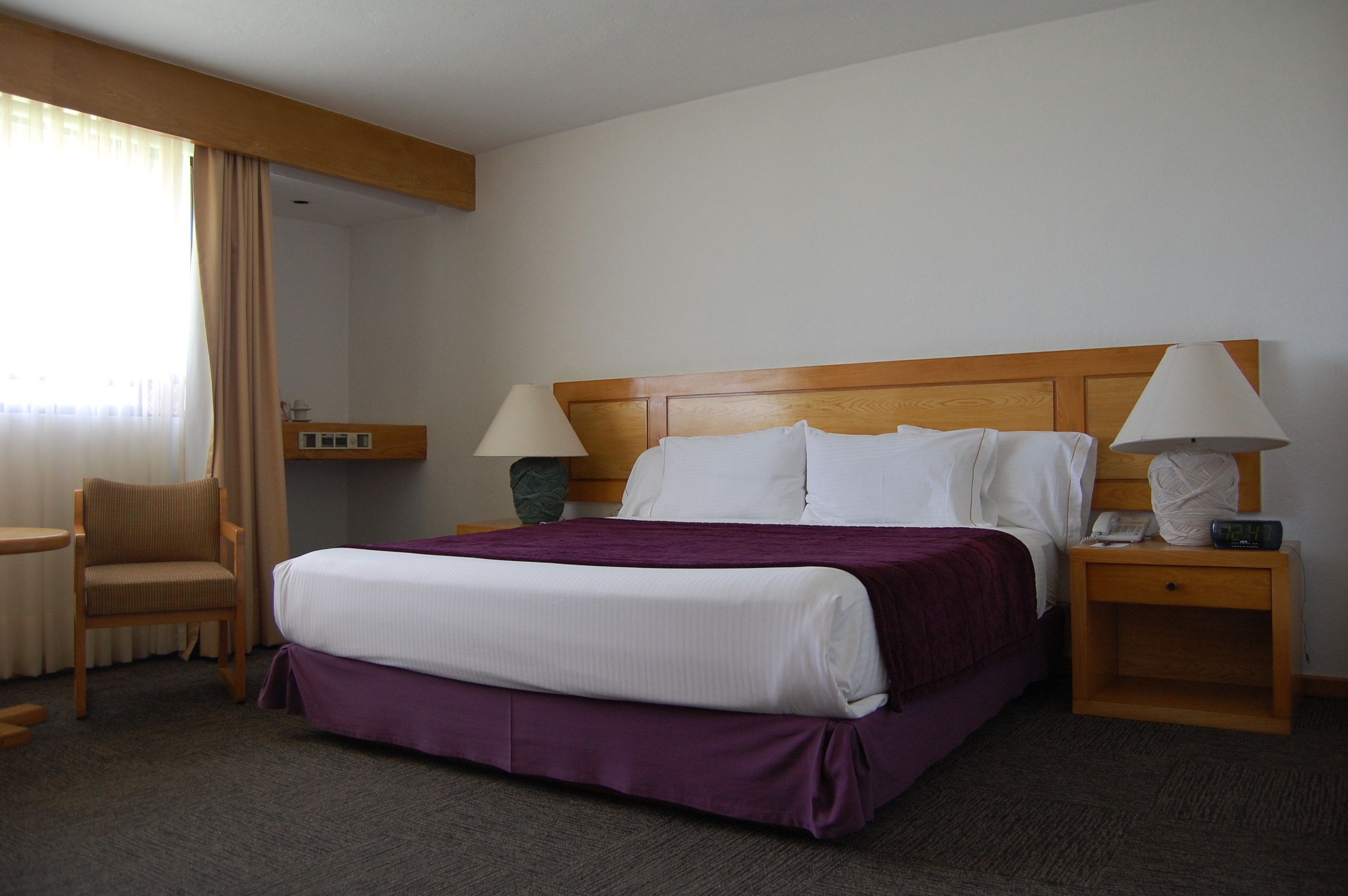 Una habitación de hotel con cama, dos mesitas de noche, una silla, una lámpara y un mueble auxiliar.