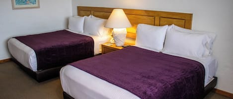 Habitación de hotel con dos camas, cada una con una colcha morada, almohadas blancas y cabeceros de madera. Hay una mesita de noche con una lámpara entre las camas.