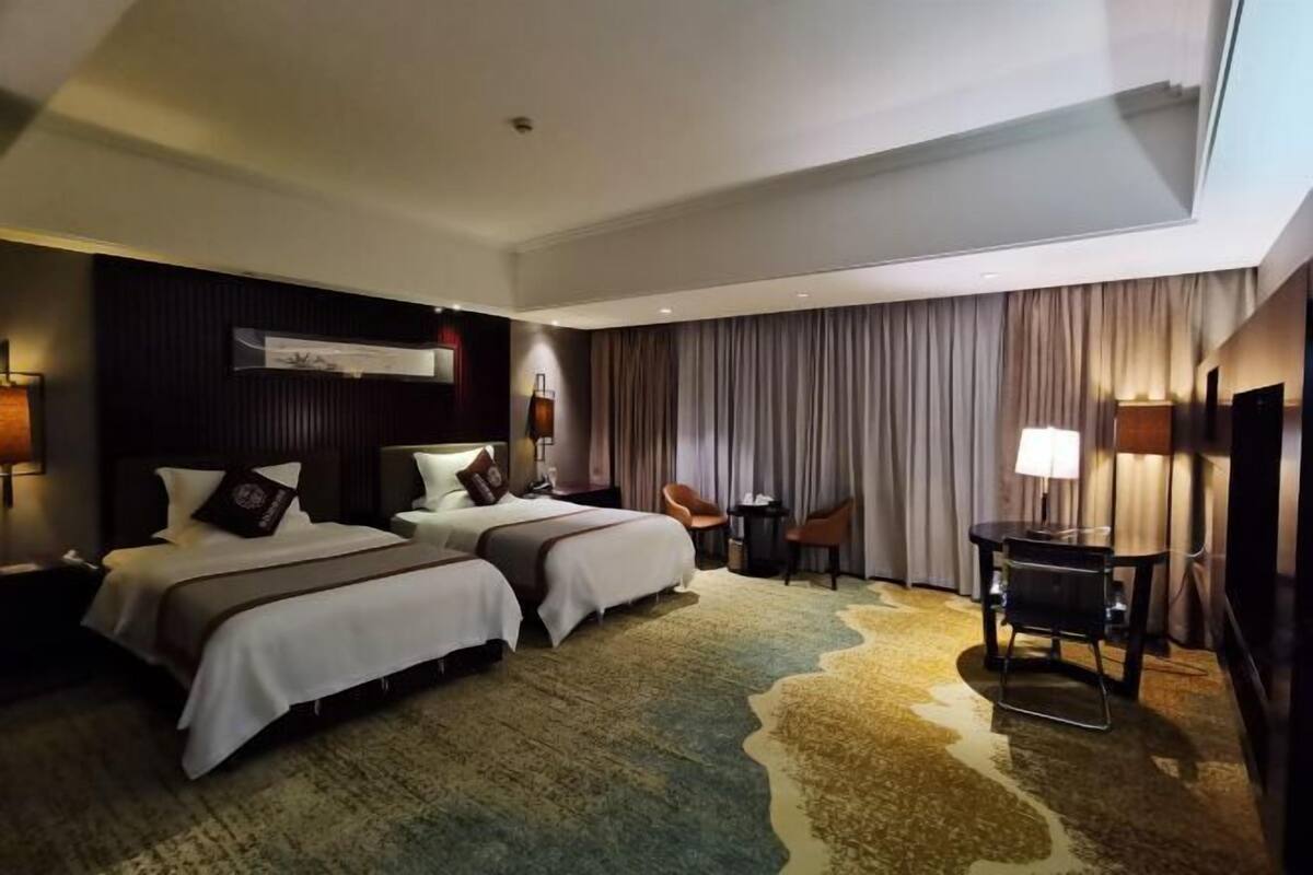 A hotel room with two beds, a desk, a chair, a TV, and a large window with curtains.