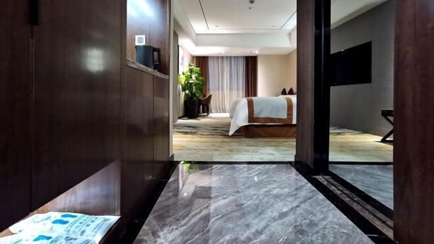 A hotel room with a marble floor, a bed with a white and brown bedspread, a plant, and a TV on the wall. - Huangting Lijing Hotel (Sanming)