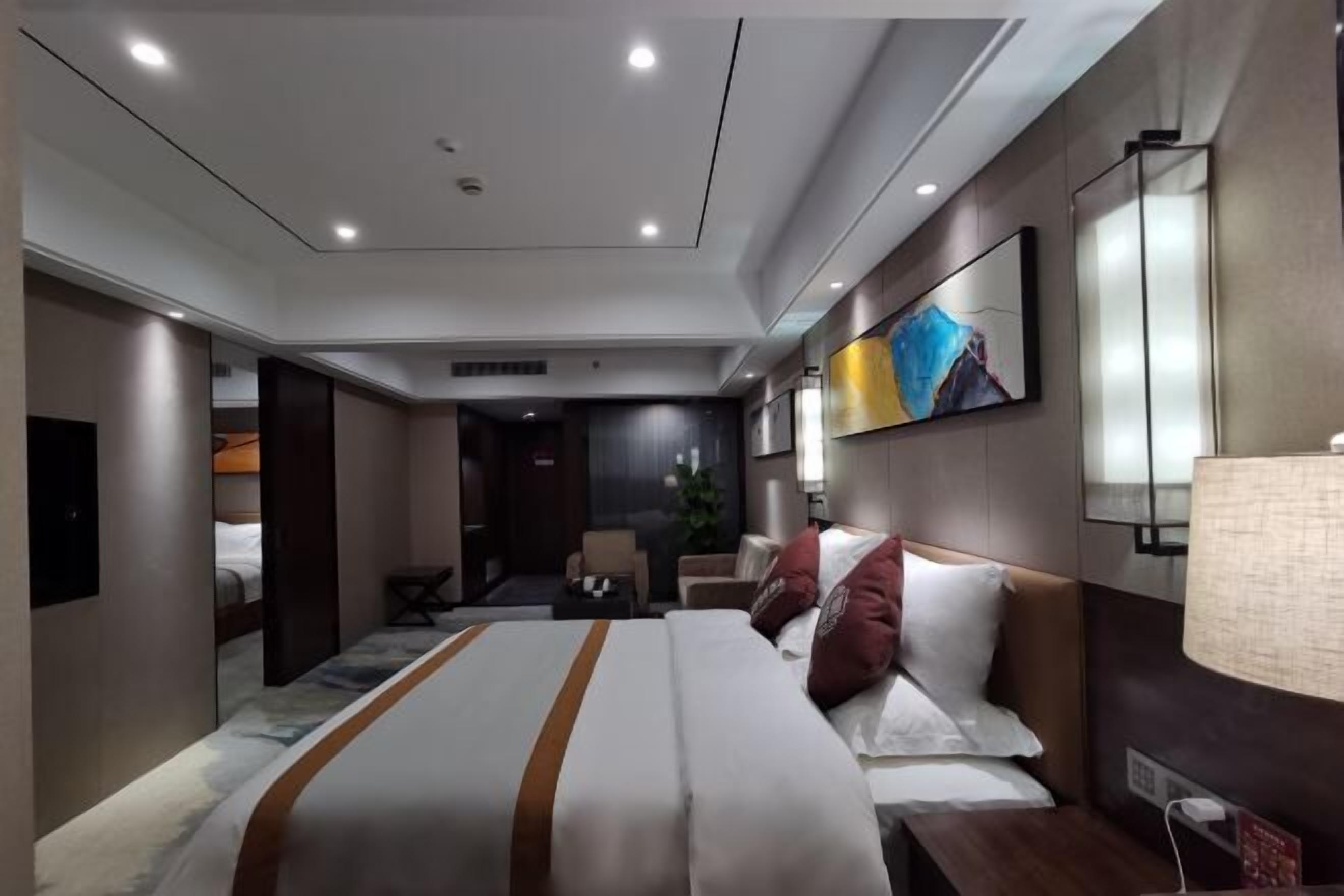A modern hotel room with a large bed, a desk, a chair, and a small table. There is a painting on the wall and a lamp on the desk.