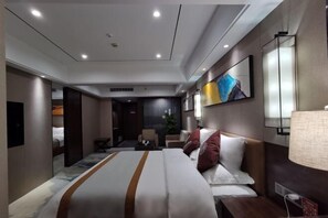 A modern hotel room with a large bed, a desk, a chair, and a small table. There is a painting on the wall and a lamp on the desk.