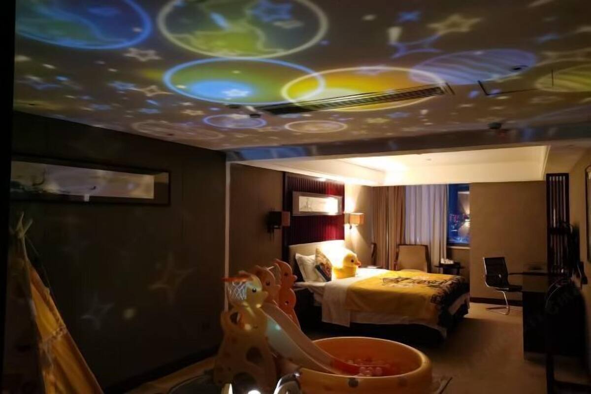 A hotel room with a bed, a desk, a chair, a projector on the ceiling, and a window.