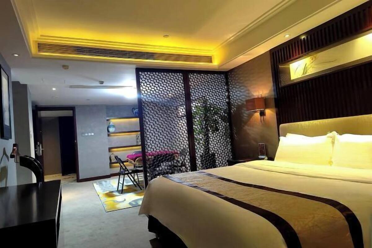 A hotel room with a large bed, a desk, and a chair. There is a partition with a decorative pattern and a plant.