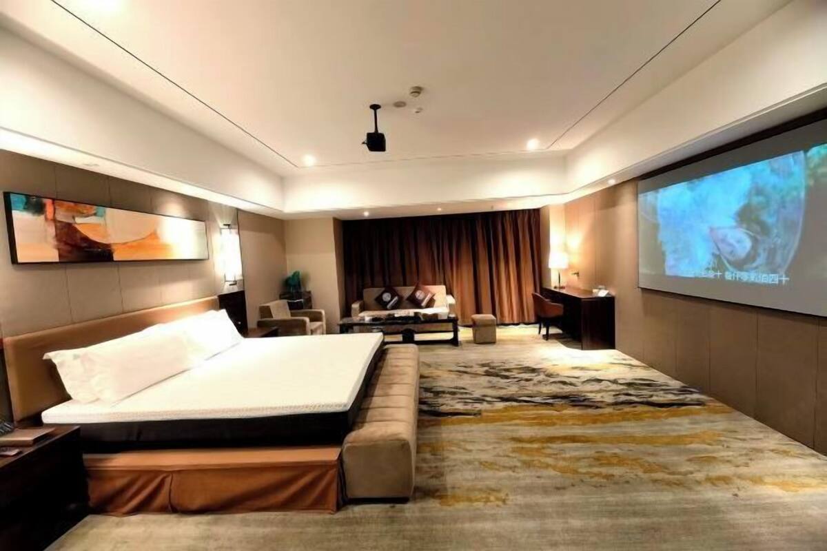 A modern hotel room with a large bed, a desk, a sofa, and a large screen displaying a movie.