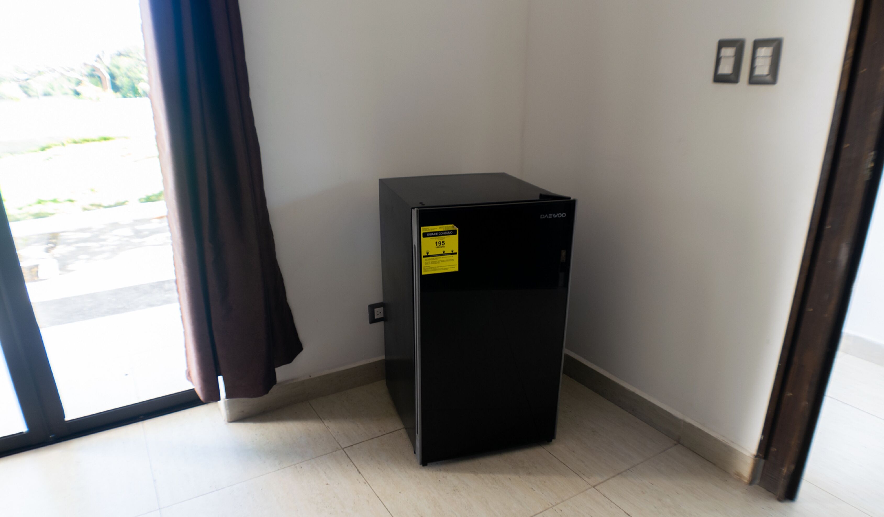 Un refrigerador negro compacto en un rincón de una habitación con piso de baldosas y una puerta que da al exterior.