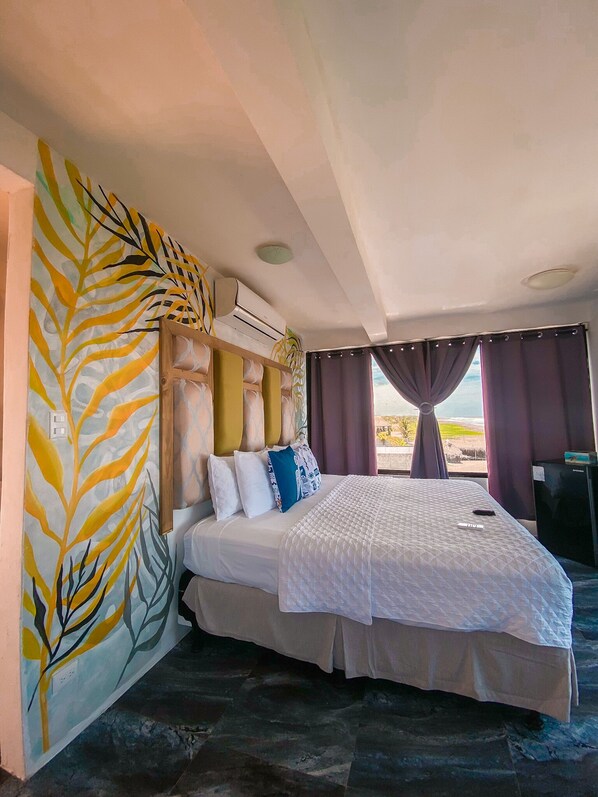 A hotel room with a large bed, a headboard with a leaf pattern, a ceiling fan, and a view of the outdoors through the window.