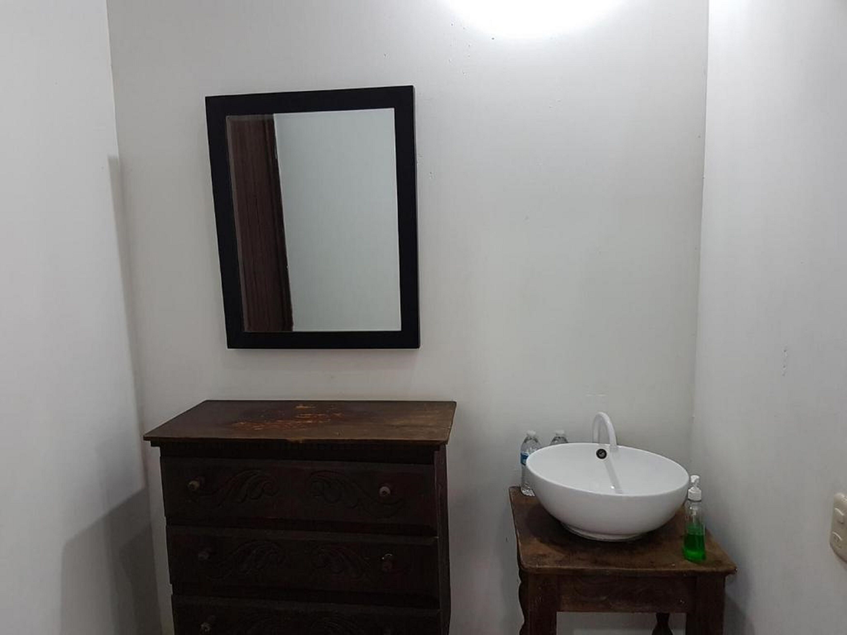 A bathroom with a wooden dresser, a white sink with a mirror above it, and a small wooden stool with a bottle of soap.