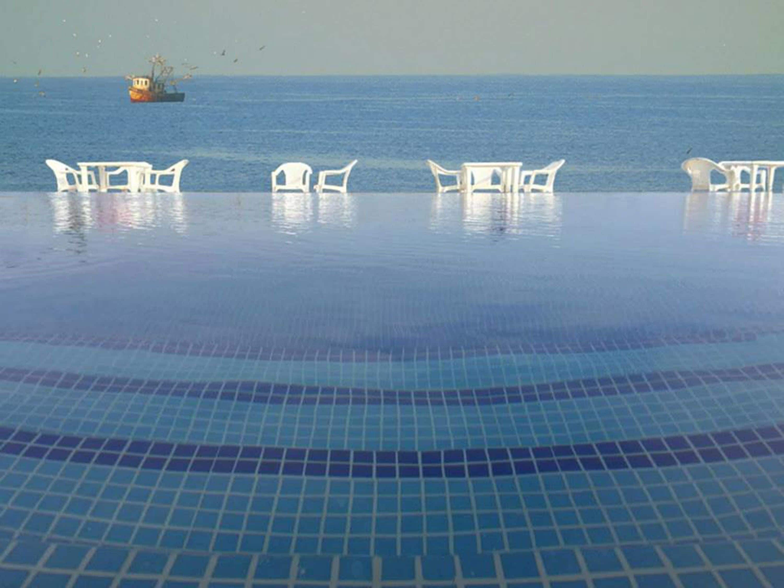 Una piscina infinita con sillas blancas y vistas al mar y un barco.