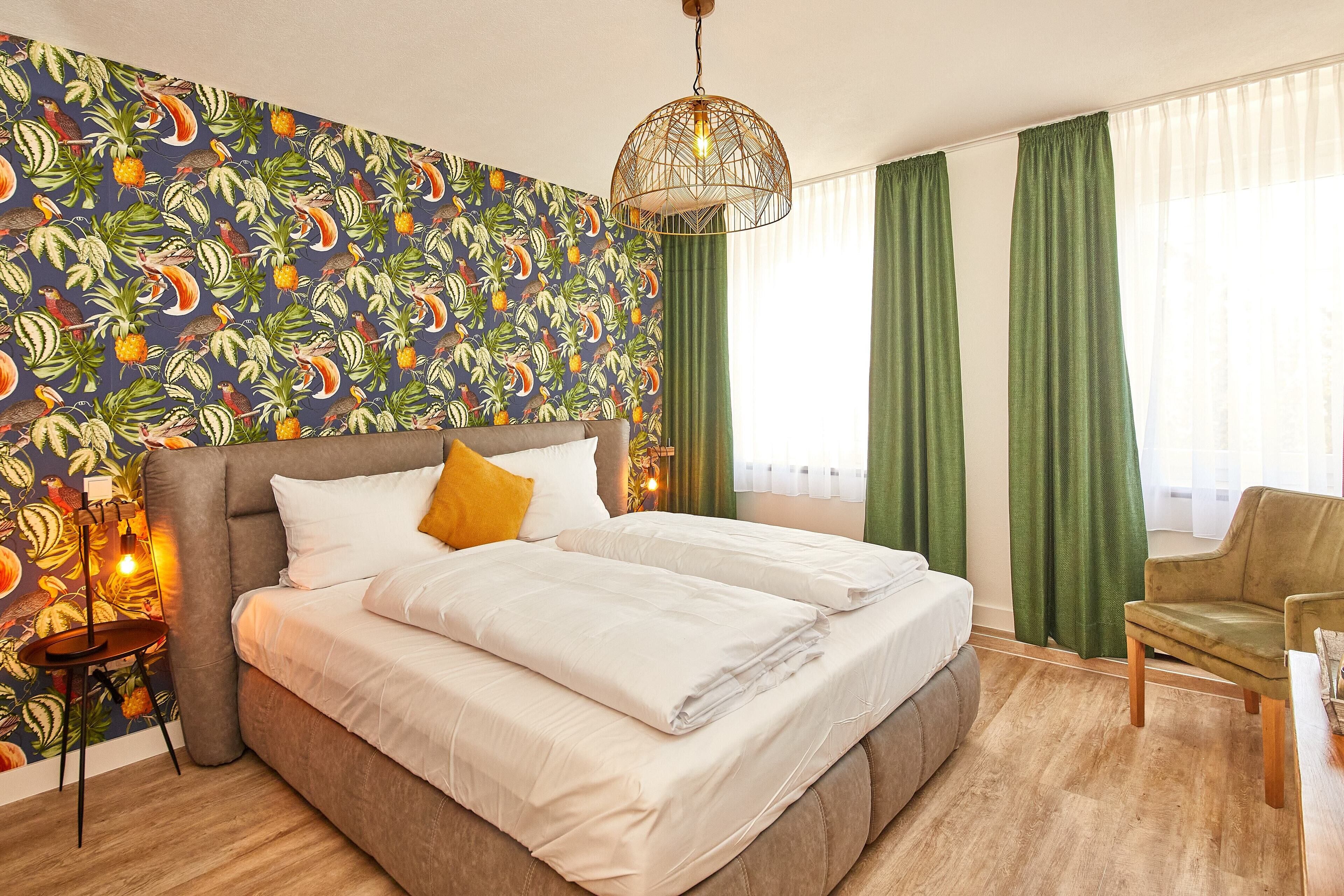 A hotel room with a large bed, a chair, a table, and a wall with a tropical wallpaper.