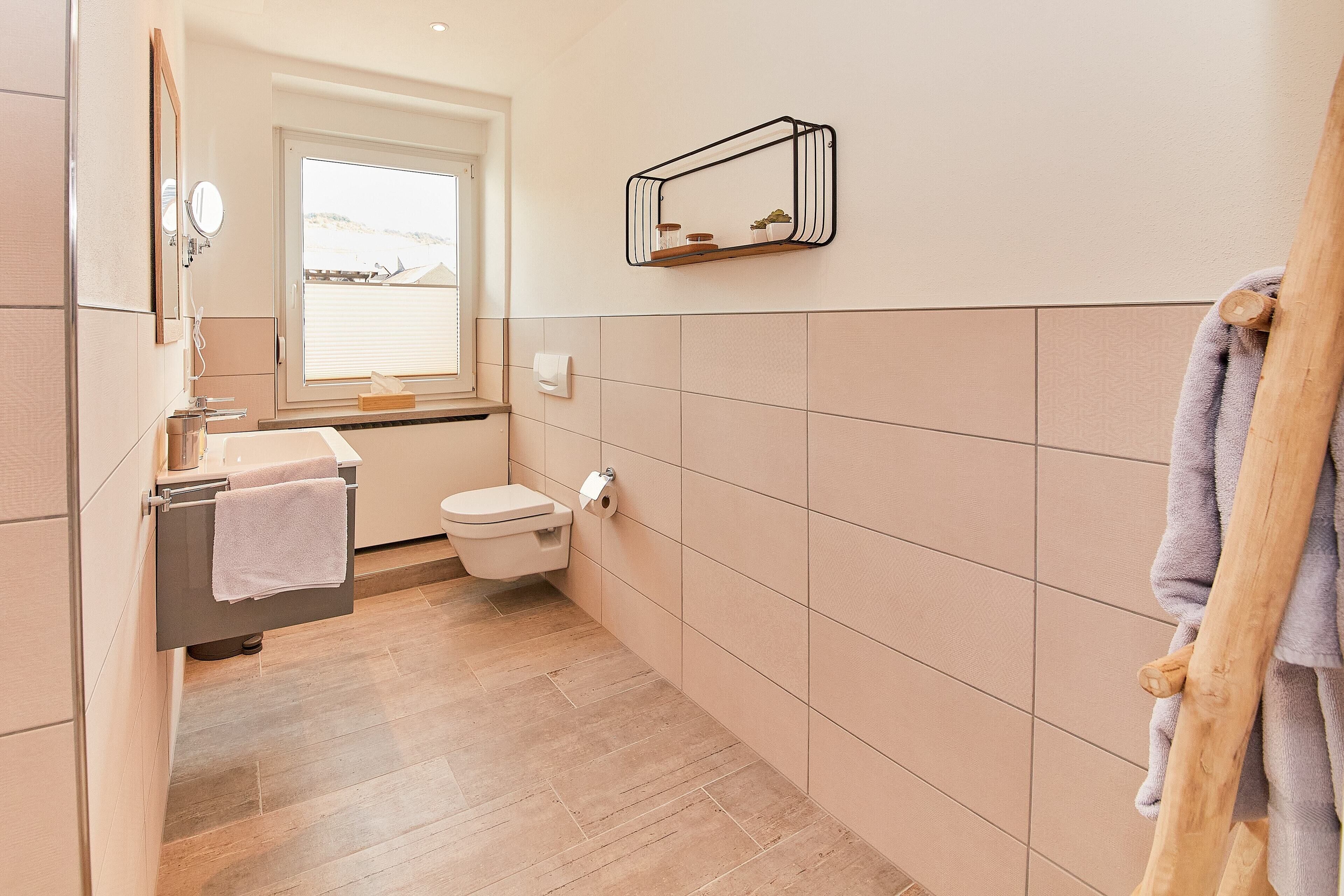 Een moderne badkamer met toilet, wastafel en spiegel. Er is een raam dat voor daglicht zorgt en een handdoekhouder met een handdoek.