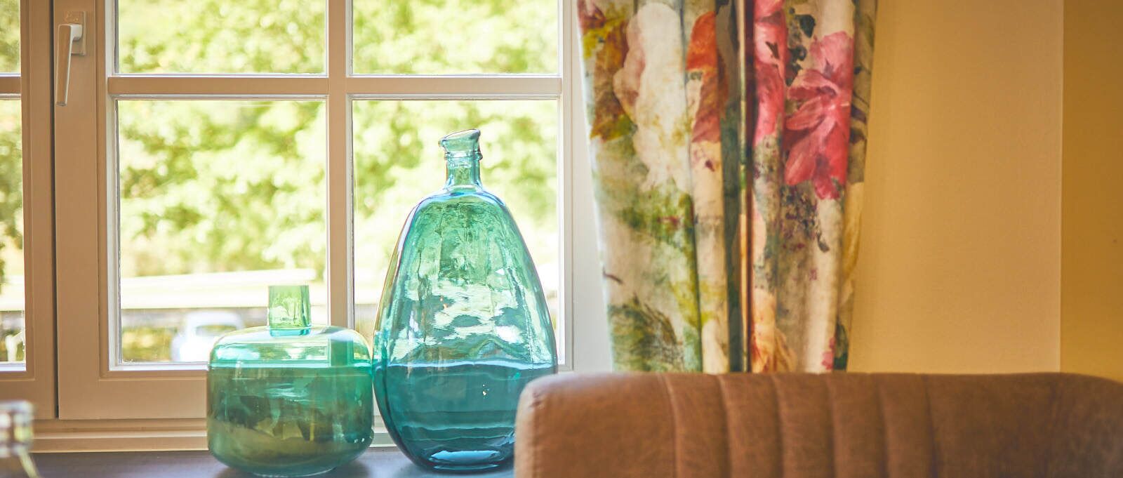 A window with two glass bottles on a windowsill, a curtain with floral patterns, and a brown chair in the foreground.