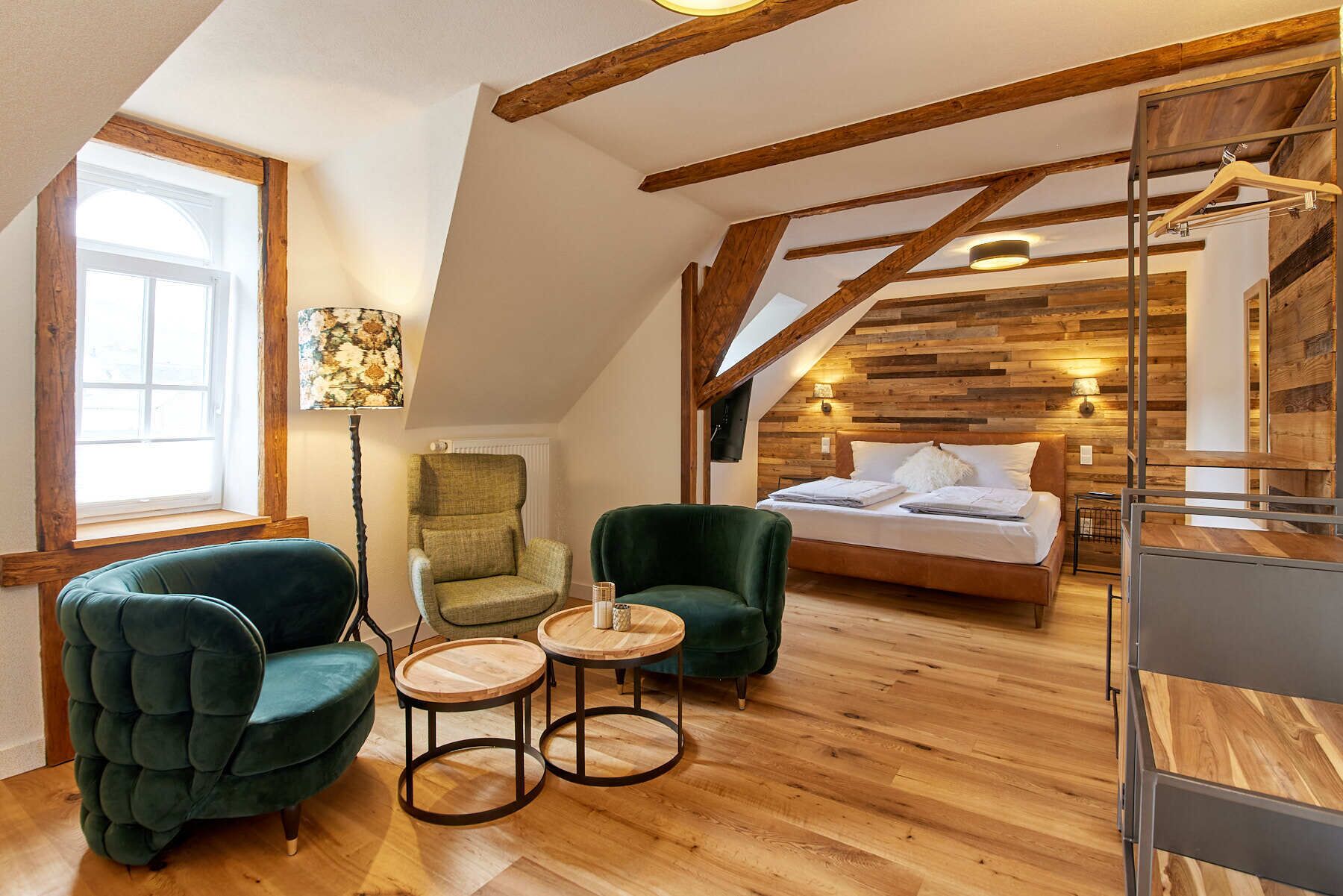 A cozy loft bedroom with a sloped ceiling, wooden beams, a bed, two armchairs, and a small table.