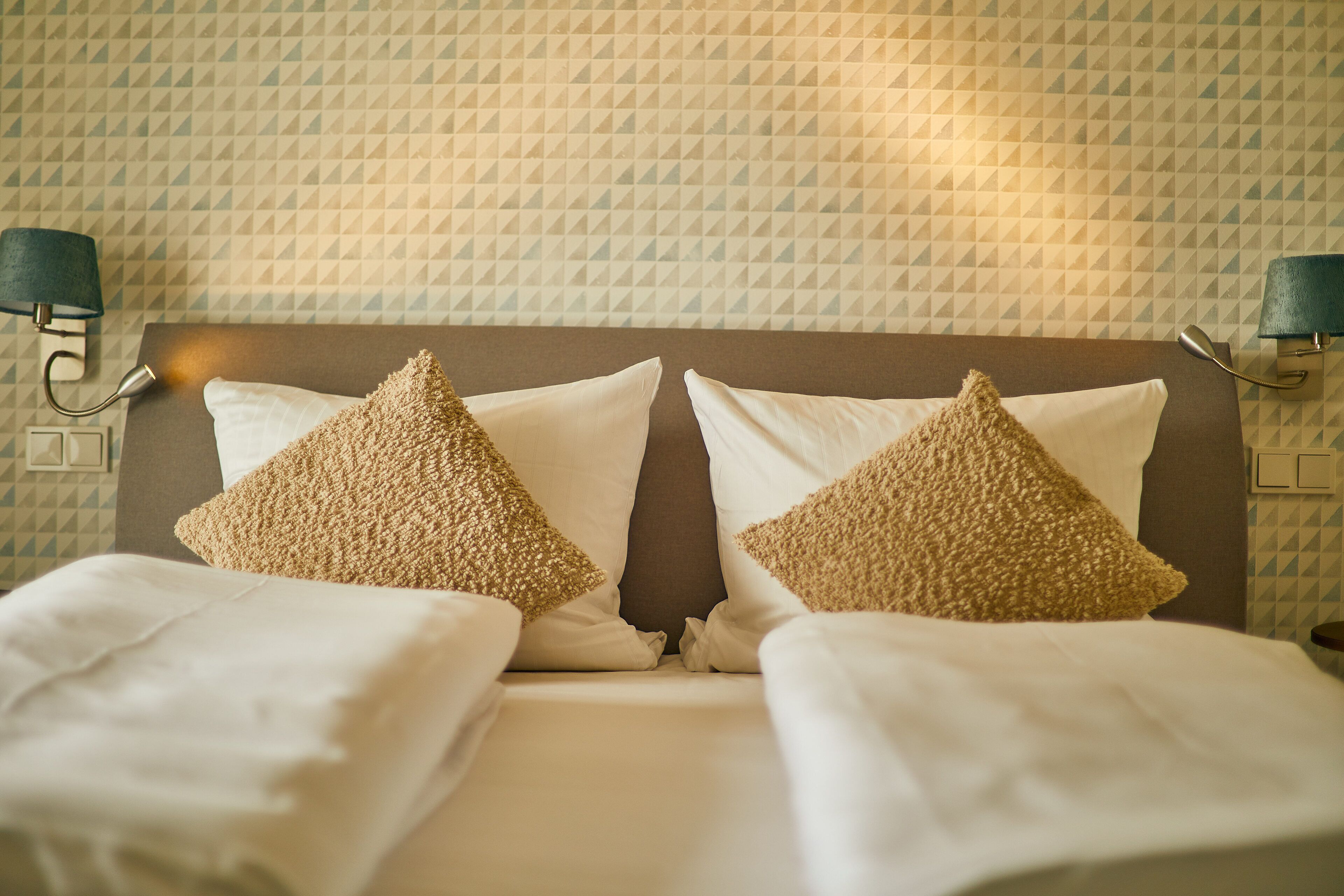 A neatly made bed with two decorative pillows and a headboard against a patterned wall.