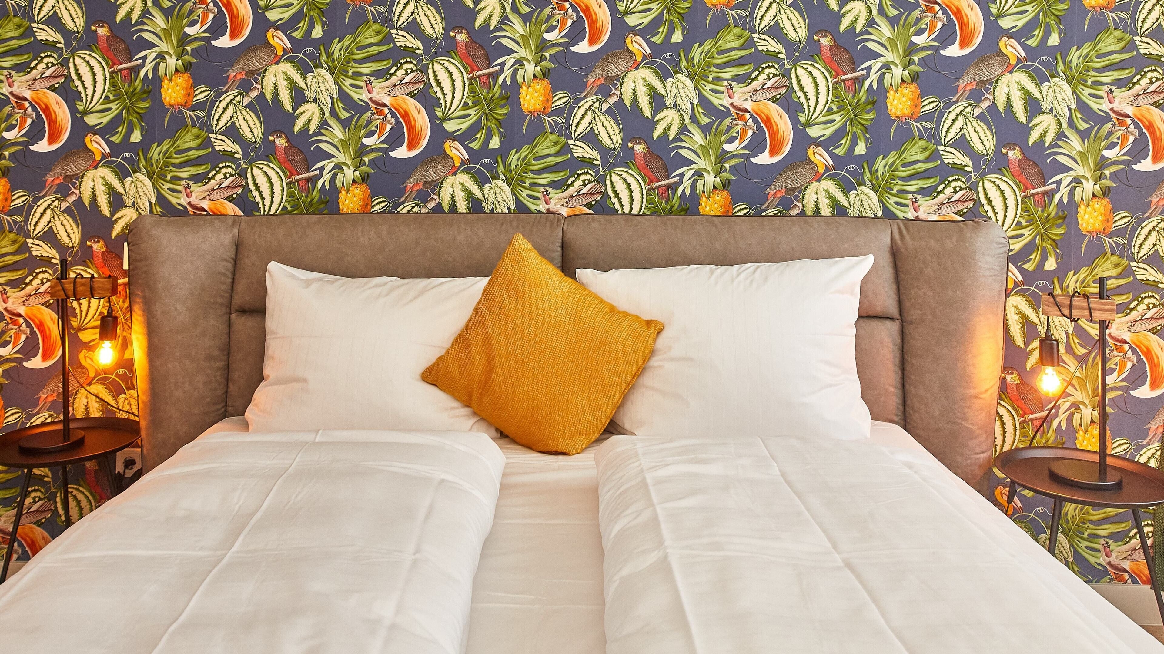 Una cama con sábanas blancas, un cojín color mostaza y un cabecero frente a una pared con estampado de aves tropicales y plantas.