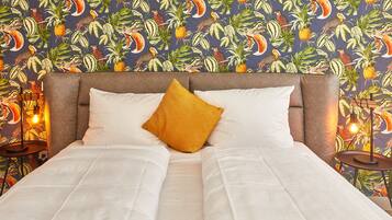Una cama con sábanas blancas, un cojín color mostaza y un cabecero frente a una pared con estampado de aves tropicales y plantas.