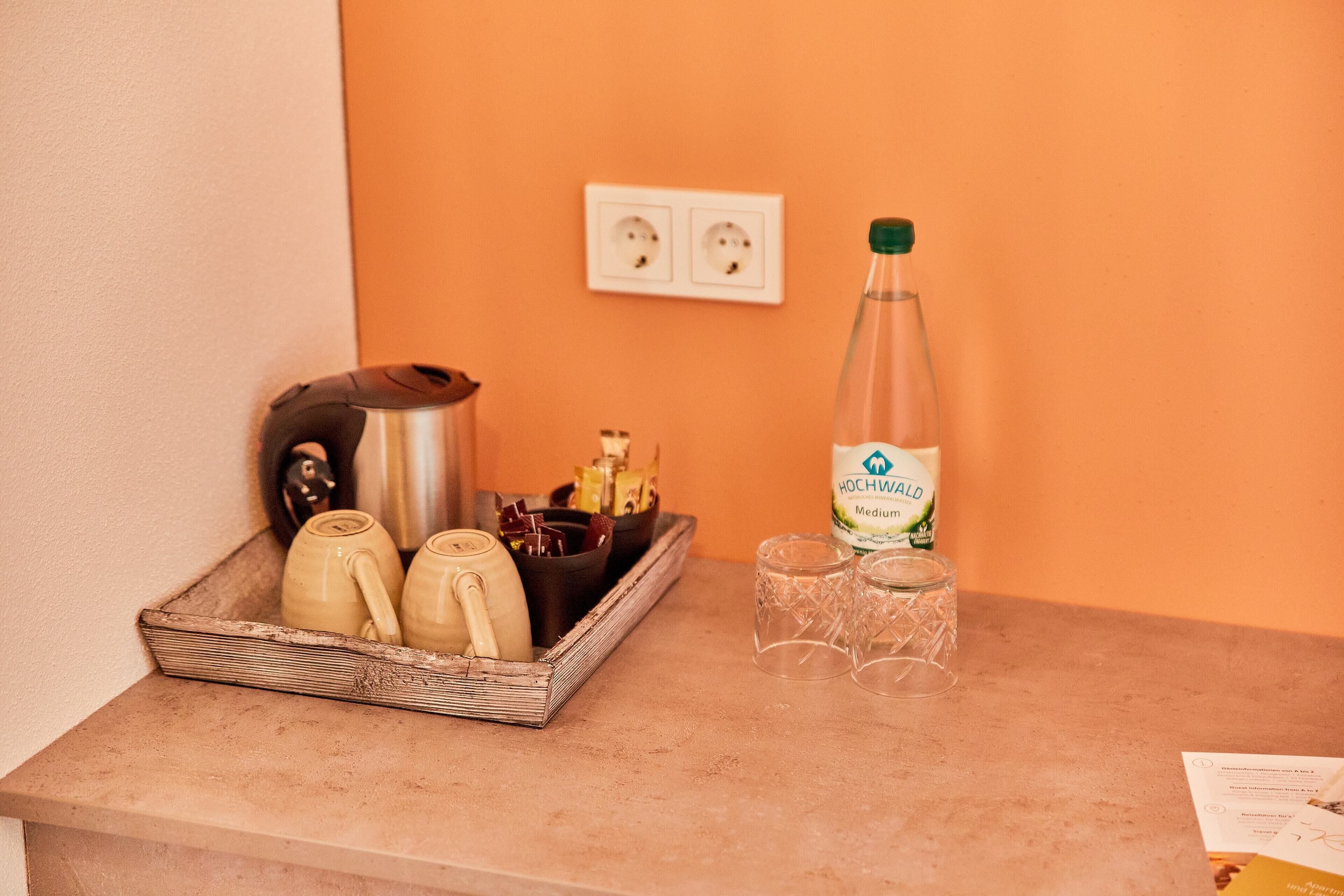 A kitchen counter with a coffee maker, teapots, a bottle of Hochwald mineral water, and two glasses.