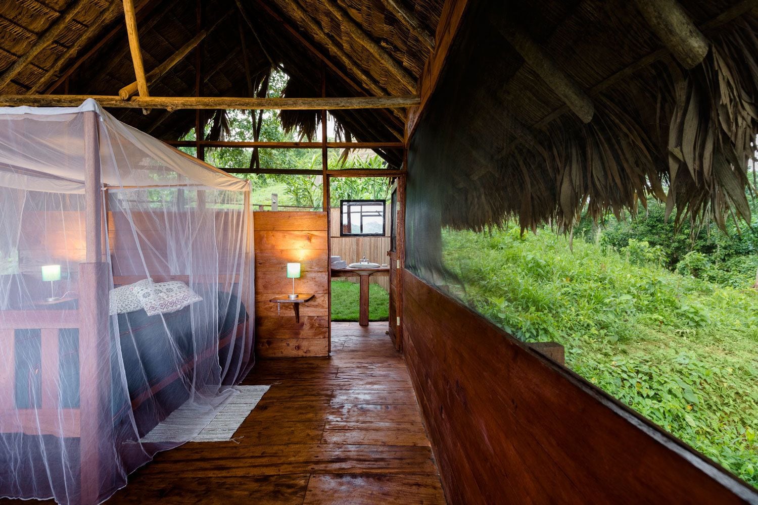 Una cabaña de madera con techo de paja, una cama con mosquitero, una mesita con lámpara y vistas a un follaje exuberante.
