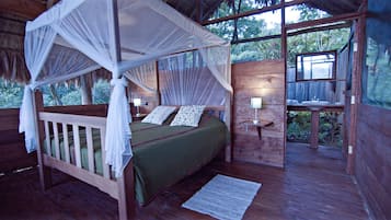 A wooden four-poster bed with a green bedspread and white pillows, a nightstand with a lamp, and a view of the surrounding forest through a window.