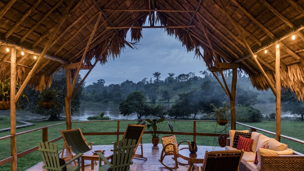 Un área de estar al aire libre con techo, mobiliario de madera y vista a un lago y bosque.