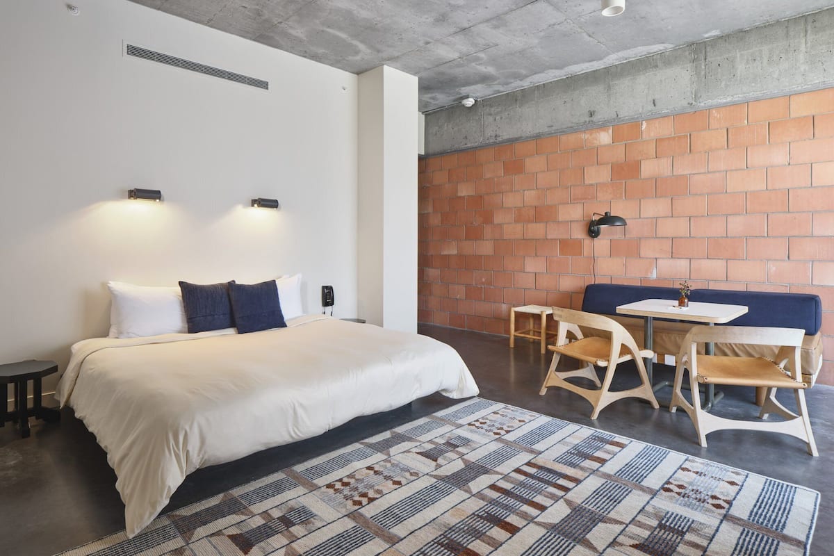 Una habitación de hotel moderna con una cama grande, una mesita, dos sillas, una pared decorada con ladrillos y una alfombra estampada.