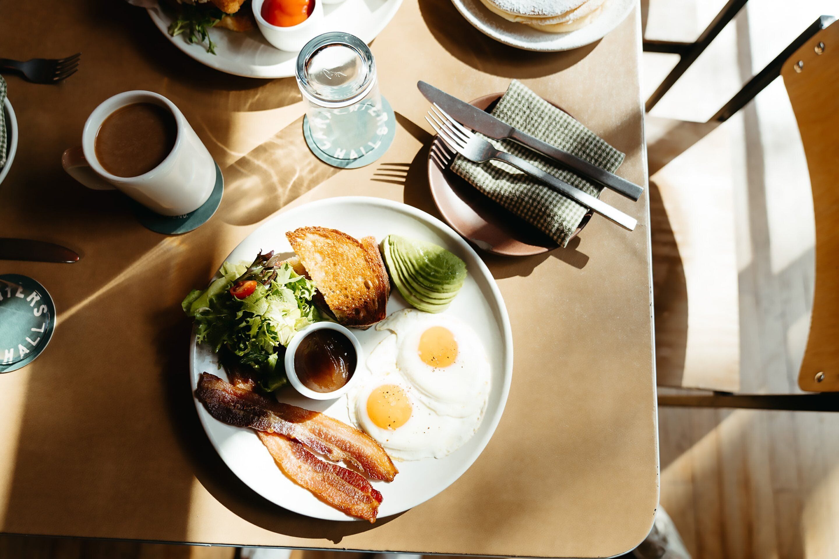 Un plato de desayuno con huevos, tocino, tostadas y ensalada sobre una mesa con una taza de café y una servilleta.