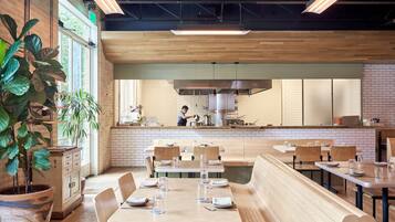A restaurant with wooden tables and benches, a kitchen in the background, and large potted plants.