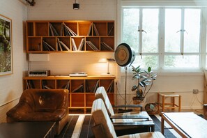 A cozy living room with a leather armchair, a sofa, a wooden bookshelf filled with books, a floor lamp, and a large window with blinds.
