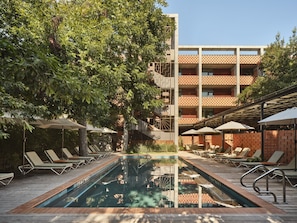 A swimming pool with lounge chairs and umbrellas, surrounded by trees and a multi-story building with a patterned facade. - The Carpenter Hotel (Austin)