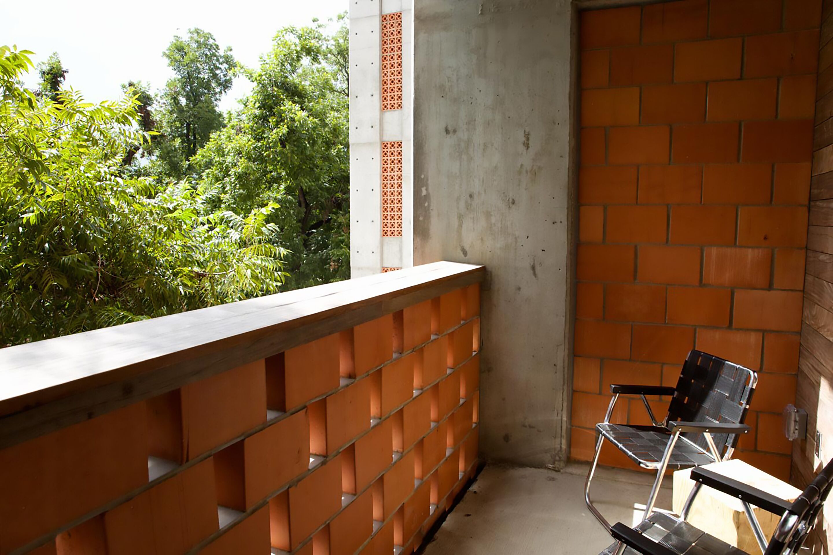 A balcony with a brick wall and a metal railing, a black chair, and a concrete pillar with a geometric pattern.