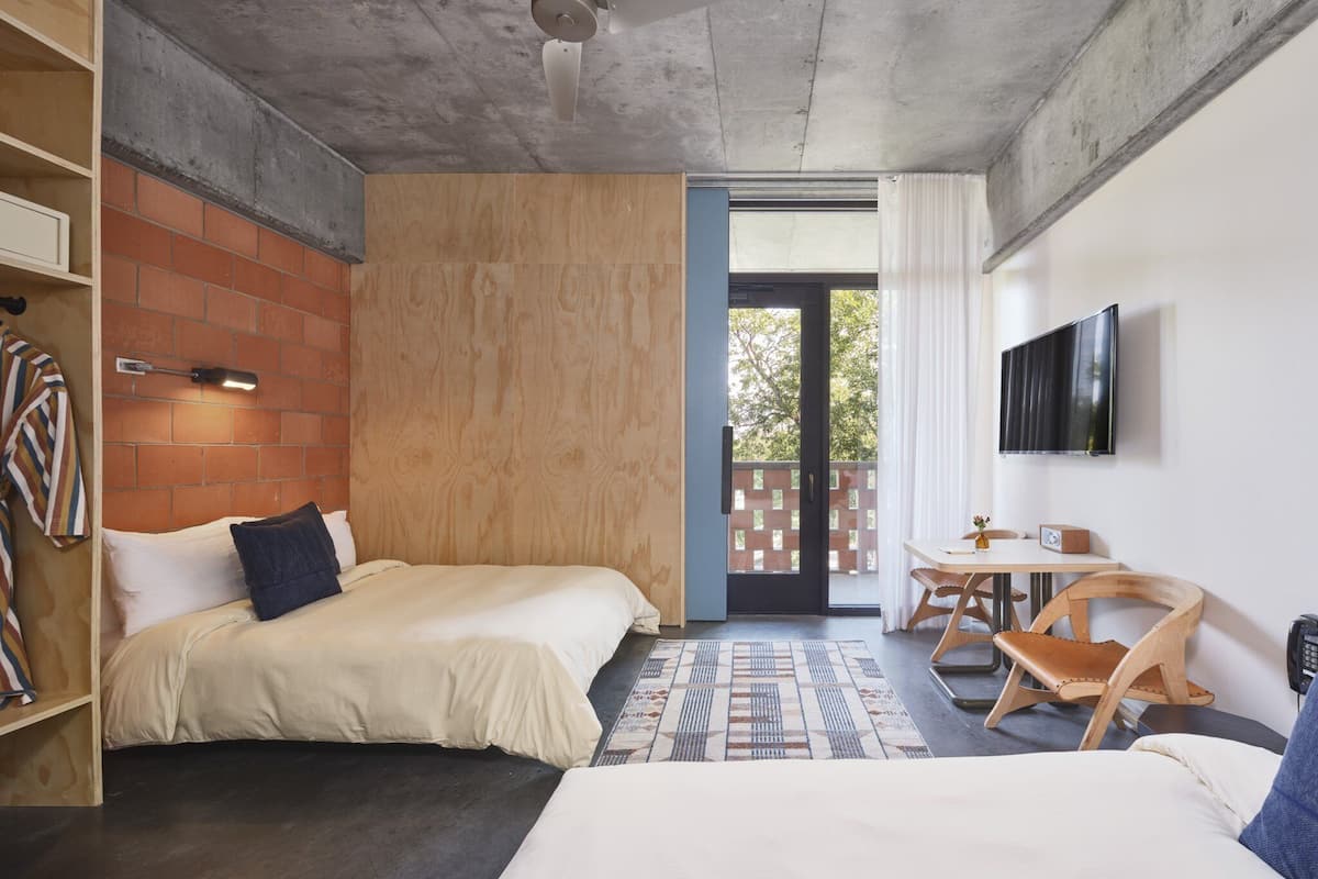 Una habitación de hotel moderna con dos camas, una pequeña mesa con sillas, televisor y un balcón con vistas.