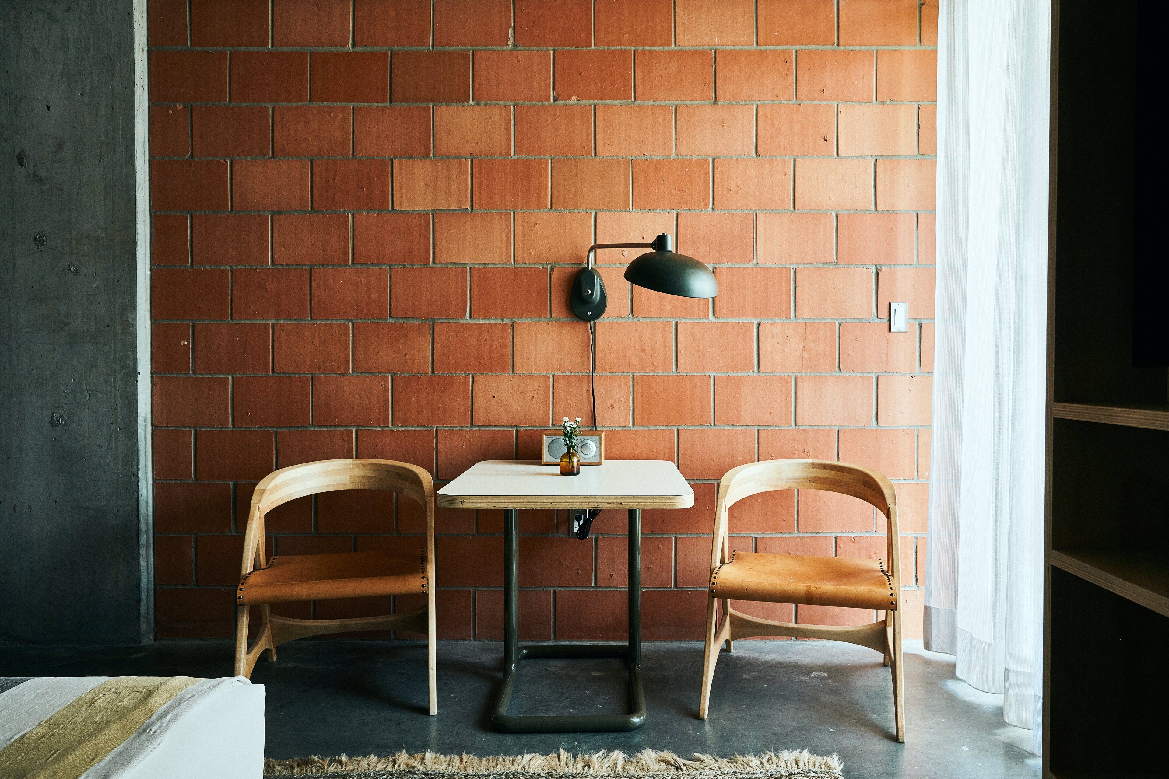 A room with a brick wall, a small table, two wooden chairs, a lamp, and a radio.