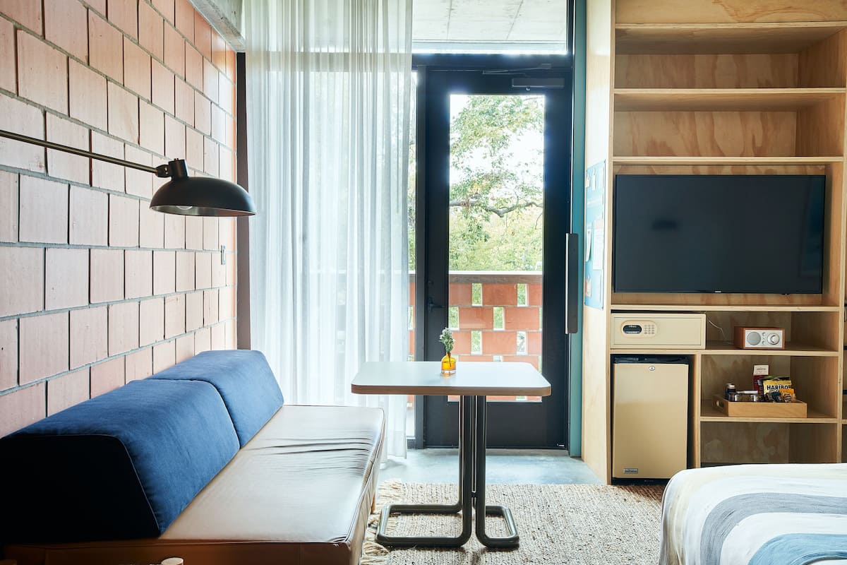 Habitación de hotel con pared de ladrillo, un sofá, una mesita con lámpara, televisión y un refrigerador pequeño.