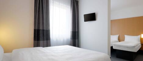 Quarto de hotel com uma cama grande, uma cama menor, televisão de tela plana e uma janela com cortinas.