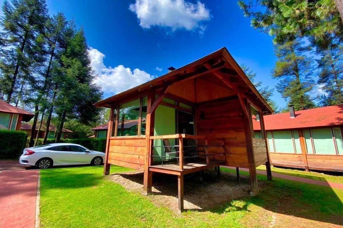 A wooden cabin with a porch, surrounded by trees and a car parked nearby.