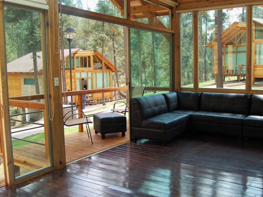 A spacious room with a large wooden sectional sofa, a dining table, and chairs. The room features large windows offering views of a forested area and a cabin.