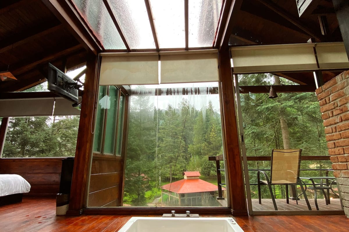 A spacious room with a large window, a wooden floor, a white bathtub, and a view of a forested area.