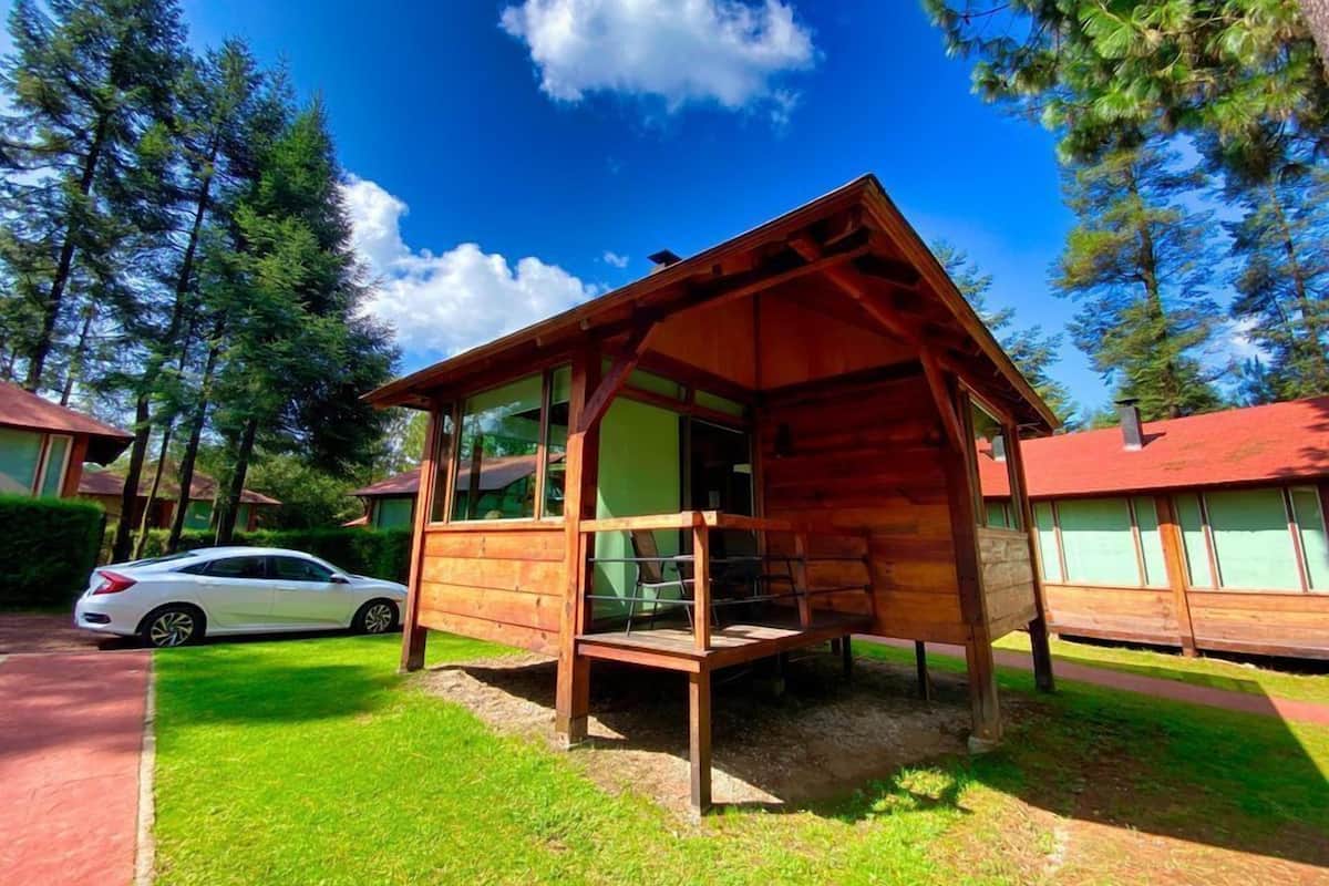 A wooden cabin with a porch, surrounded by trees and a white car parked nearby.