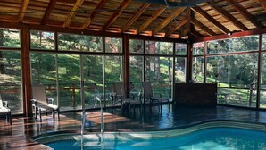 A large indoor swimming pool with a wooden deck and seating area, surrounded by large windows offering views of the outdoors. - Quinta Los Azufres (Ciudad Hidalgo)
