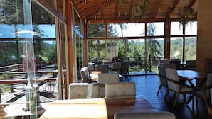 An outdoor dining area with wooden tables and chairs, large windows offering views of the forest, and hanging plants. - Quinta Los Azufres (Ciudad Hidalgo)