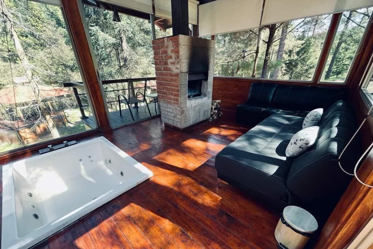 A room with a fireplace, a black sofa, a white bathtub, and large windows overlooking a forest.