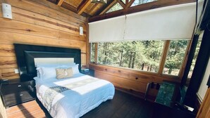 A wooden cabin bedroom with a bed, a nightstand, and a large window offering a forest view. - Quinta Los Azufres (Ciudad Hidalgo)