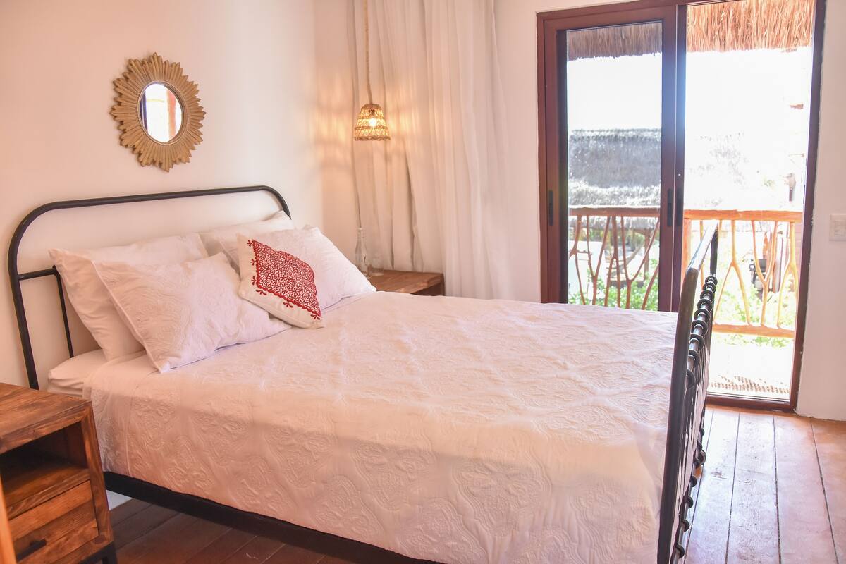 Una cama con ropa de cama blanca y una almohada roja, una mesita de noche de madera, un espejo en la pared y vistas al exterior a través de una puerta corrediza de vidrio.
