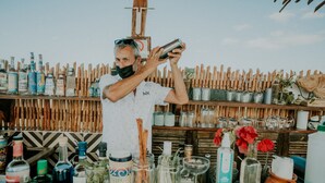 A bar area with a variety of bottles and jars on shelves, a wooden fence in the background, and a person preparing a drink. - Hotel Casa HX - Adults Only (Isla Holbox)