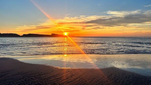 Una playa con un atardecer sobre el océano, olas tranquilas y un cielo despejado.