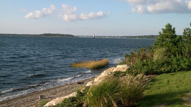 A coastal landscape with a rocky shoreline, a lighthouse in the distance, and a grassy area with vegetation. - Waterfront - secluded beachfront 
Last Min Discount for week of August 29-Sept 5 (Portsmouth)