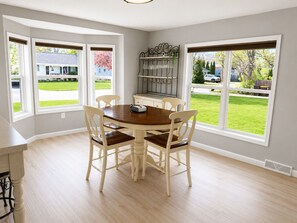 Un coin repas comprenant une table ronde, quatre chaises et de grandes fenêtres donnant sur une pelouse et des maisons.