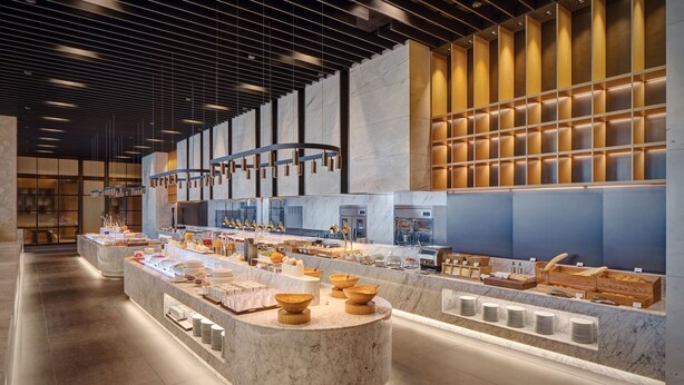 Un hall d’hôtel moderne et bien éclairé, avec un comptoir d’accueil en marbre, divers points de vente de nourriture et de boissons, et un grand espace salon.